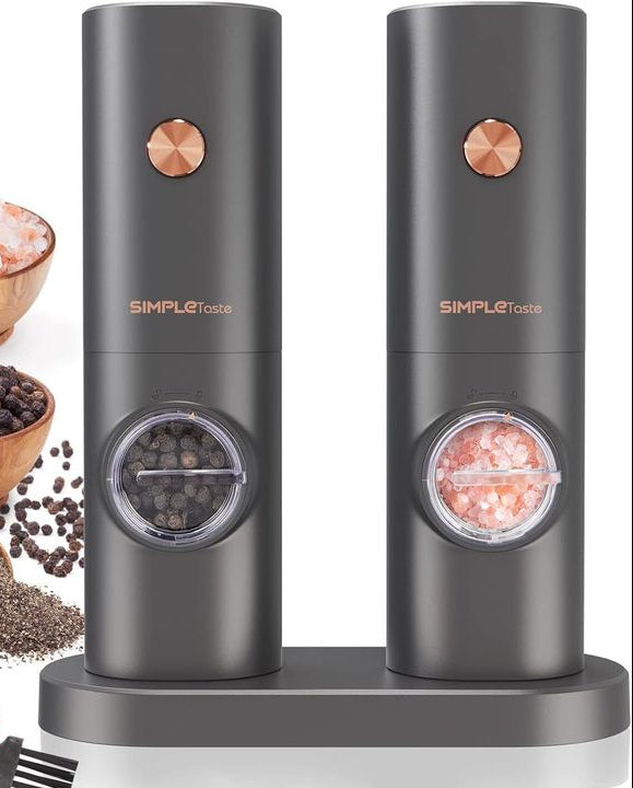 Two black pepper grinders with copper accents on a white background, surrounded by spices and a power cord.